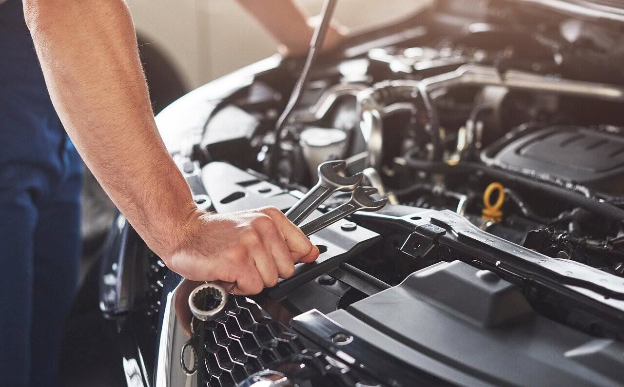 A mechanic is working on a vehicle engine. In his hand, he holds a wrench.