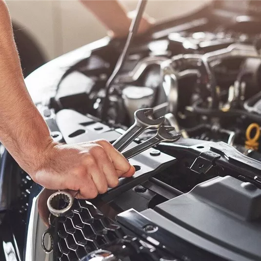 A mechanic is working on a vehicle engine. In his hand, he holds a wrench.