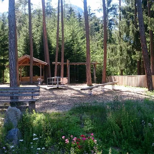 A quiet forest spot with a wood shelter and a cabin. Meadows are blossoming with flowers all around, and tall trees provide shade.