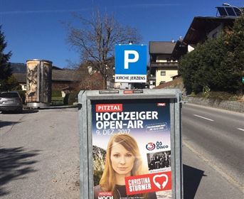Ein Plakat an einem Ständer für das Hochzeiger Open-Air am 9. Dezember 2017. Im Hintergrund sind eine Straße und einige Gebäude zu sehen.