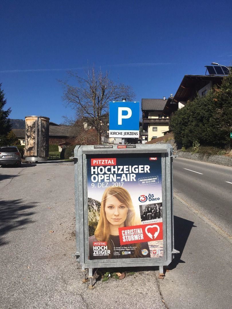 Ein Plakat für das Hochzeiger Open-Air Event, das am 9. Dezember 2017 stattfindet. Im Hintergrund ist eine Parkplatzeinfahrt und eine ruhige Straße zu sehen.