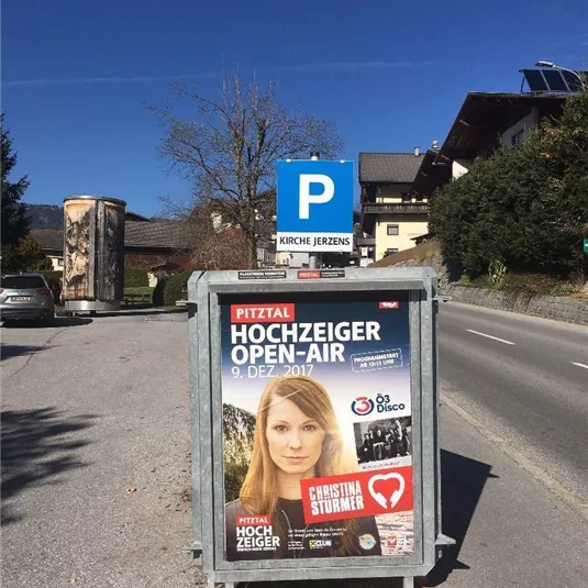 Ein Plakat für das Hochzeiger Open-Air Event, das am 9. Dezember 2017 stattfindet. Im Hintergrund ist eine Parkplatzeinfahrt und eine ruhige Straße zu sehen.