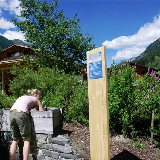 A person is washing their hands at a water basin in a natural setting. In the background, green hills and traditional wooden houses can be seen.