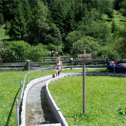 A green field with a stream and a sign indicating a recreation area. In the background, there are trees and some people relaxing.