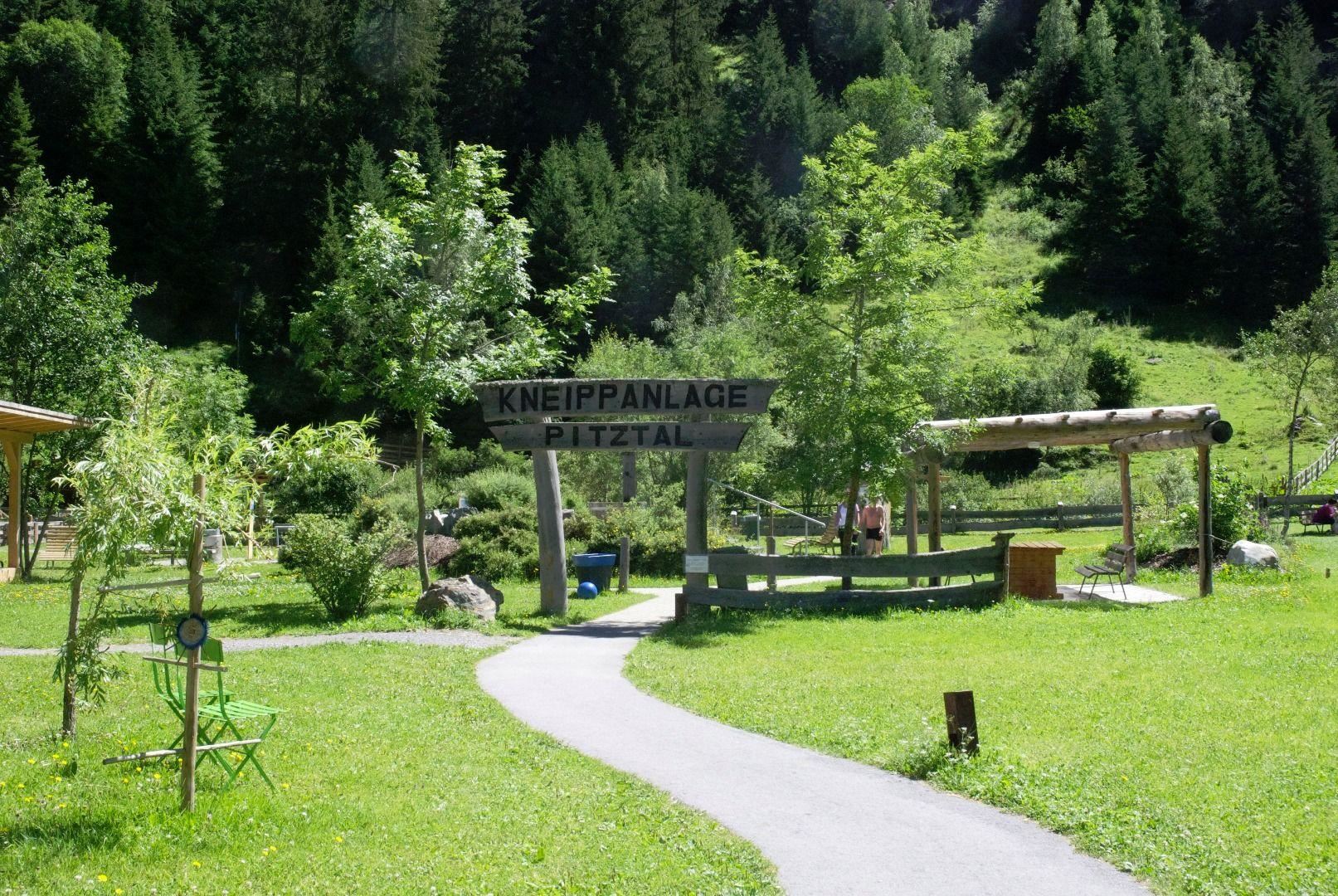 A green meadow with a path and trees. In the foreground stands a sign that reads "Kneippanlage Pitztal".