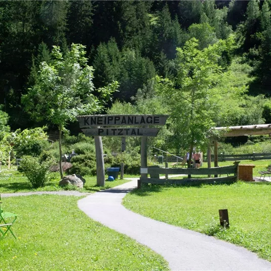 A green meadow with a path and trees. In the foreground stands a sign that reads "Kneippanlage Pitztal".
