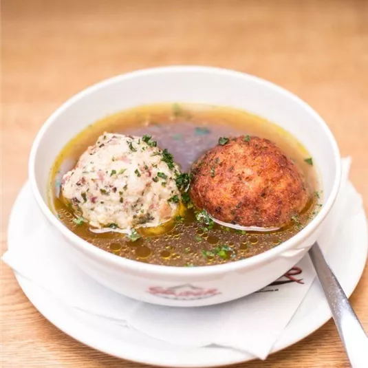 A bowl of broth containing two dumplings. The clear broth is garnished with fresh herbs.