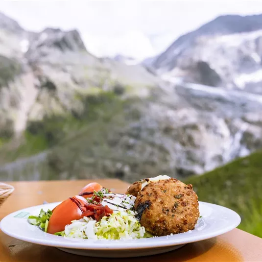 Ein Teller mit gebratenem Gericht, Salat und Tomaten steht auf einem Tisch im Freien. Im Hintergrund sind beeindruckende Berge und eine grüne Landschaft zu sehen.