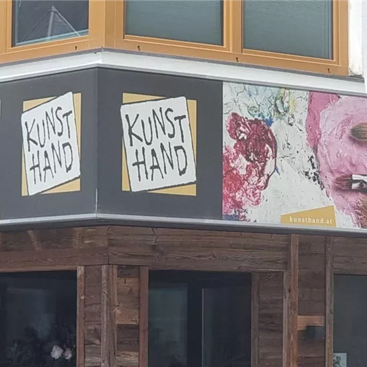 Ein modernes Geschäftsschild mit dem Namen "KUNST HAND". Das Schild zeigt eine bunte Grafik von Malutensilien und Kunstwerken.