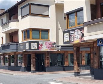 Ein modernes Gebäude mit mehreren Balkonen und Schaufenstern in einer ruhigen Straße. Im Hintergrund sind Bäume und ein klarer Himmel sichtbar.