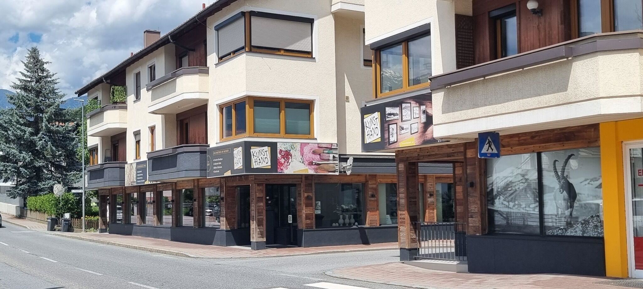 Ein modernes Gebäude mit mehreren Balkonen und Schaufenstern in einer ruhigen Straße. Im Hintergrund sind Bäume und ein klarer Himmel sichtbar.