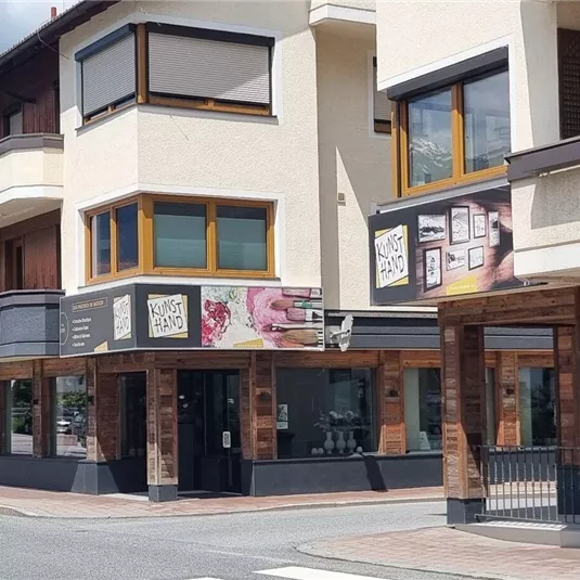 Ein modernes Gebäude mit mehreren Balkonen und Schaufenstern in einer ruhigen Straße. Im Hintergrund sind Bäume und ein klarer Himmel sichtbar.