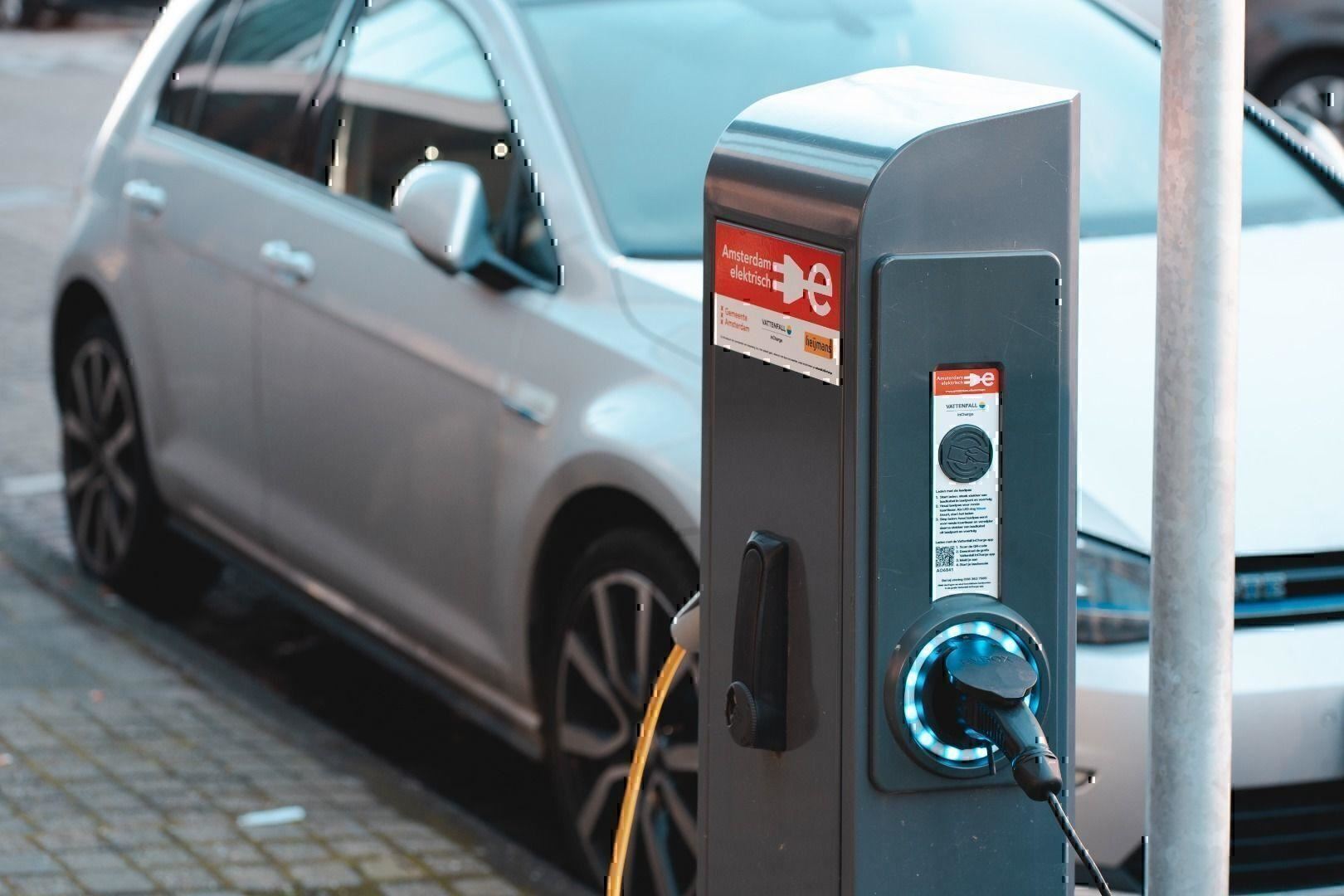 A charging station for electric cars stands by the street, while a silver car is parked in the background. The cables are connected to the station.
