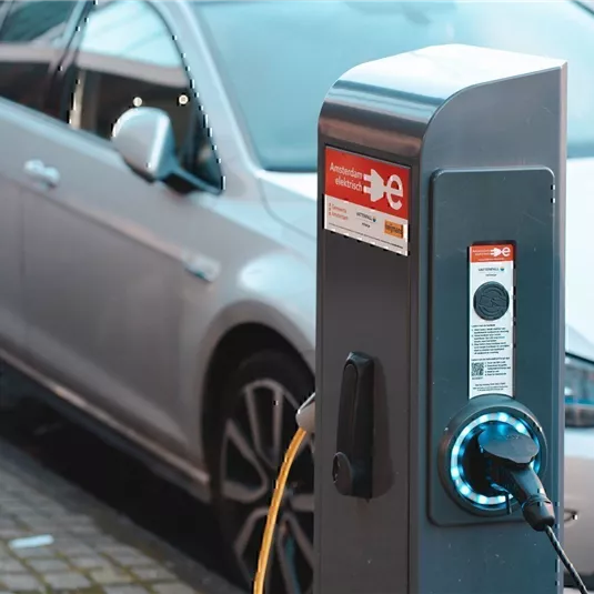 Eine Ladestation für Elektrofahrzeuge steht an der Straße. Im Hintergrund ist ein silbernes Auto geparkt.