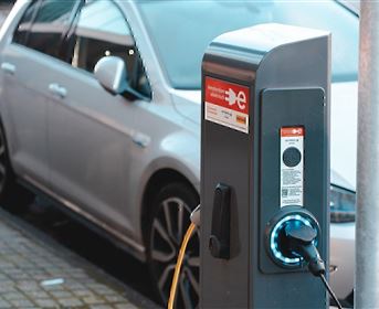 Eine Ladestation für Elektroautos mit einem Kabel, das an ein Fahrzeug angeschlossen ist. Im Hintergrund steht ein silbernes Auto.