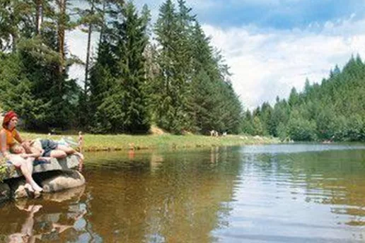 Ein ruhiger See umgeben von hohen Bäumen und grünen Wiesen. Zwei Personen entspannen am Ufer und genießen die Natur.