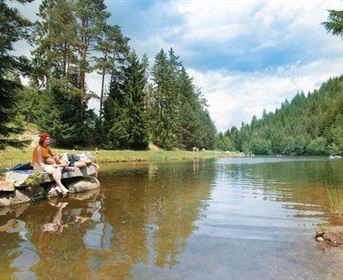 Ein ruhiger See umgeben von hohen Bäumen und grünen Wiesen. Zwei Personen entspannen am Ufer und genießen die Natur.