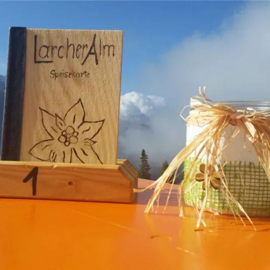 A wooden menu book is on a table with a mountain view. Next to it is a decorative glass with a ribbon.