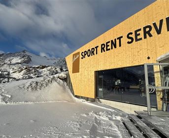 A modern wooden building with the inscription "SPORT RENT SERVICE" is located in the snow. In the background, mountains and a clear sky can be seen.