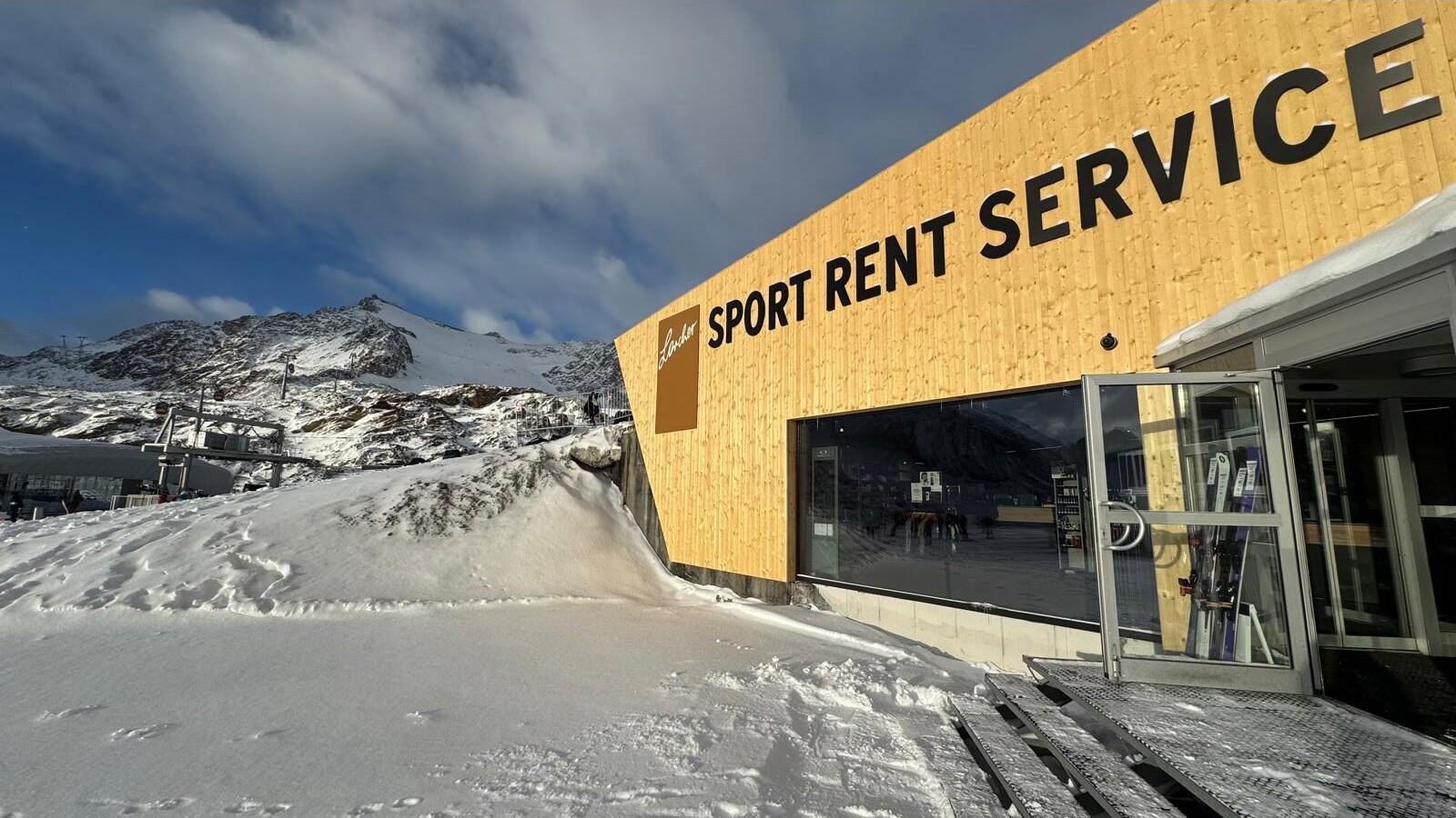Ein modernes Holzgebäude mit der Aufschrift "SPORT RENT SERVICE" liegt im Schnee. Im Hintergrund sind Berge und ein klarer Himmel zu sehen.