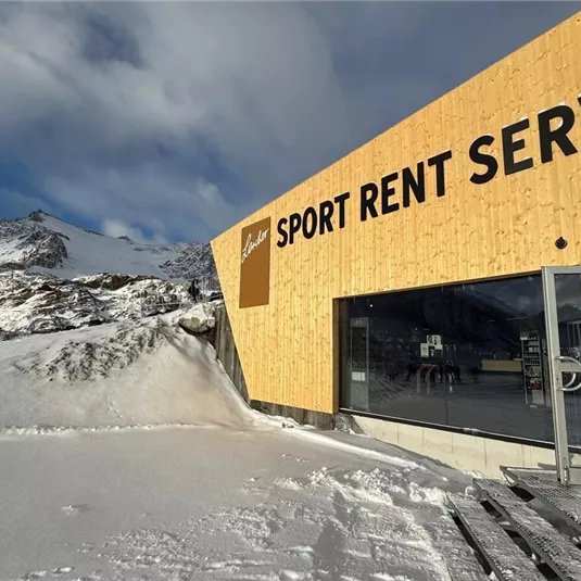 Ein modernes Holzgebäude mit der Aufschrift "SPORT RENT SERVICE" liegt im Schnee. Im Hintergrund sind Berge und ein klarer Himmel zu sehen.
