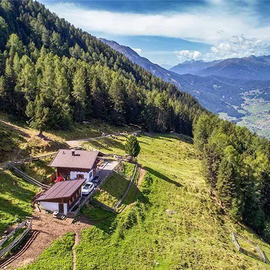 Ein malerisches Grundstück in den Bergen mit einer kleinen Hütte umgeben von Bäumen. Im Hintergrund sind die Berge und das Tal sichtbar.