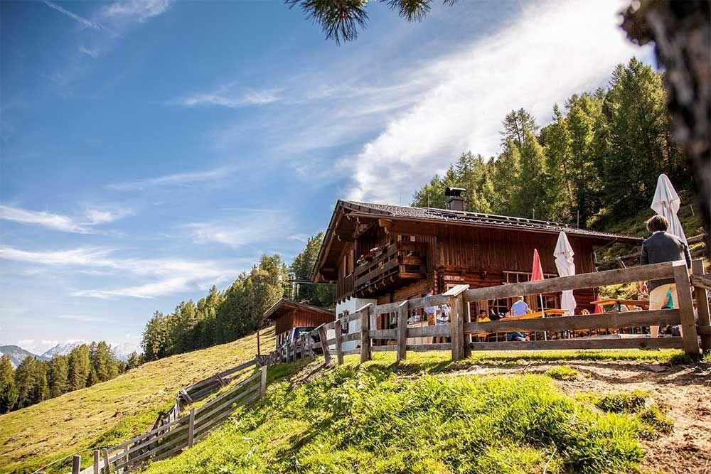 Eine malerische Berghütte umgeben von Wiesen und Bäumen. Der Himmel ist klar und blau, was eine einladende Atmosphäre schafft.