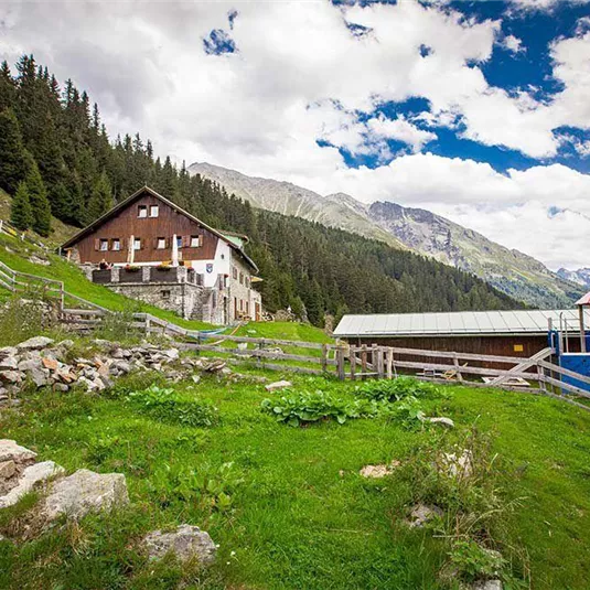An alpine landscape with several huts and green meadows. In the background, mountains and a cloudy sky are visible.