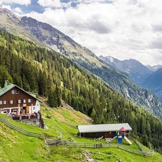 A beautiful mountain cabin surrounded by many trees and gentle hills. In the background, impressive mountains and a clear sky can be seen.