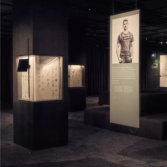 Ein moderner Ausstellungsraum mit mehreren Vitrinen und Informationen an Wänden. Ein großes Bild einer Person ist in der Mitte des Raumes sichtbar.