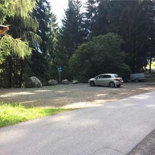 Ein Parkplatz in einem Waldgebiet mit mehreren Bäumen. Ein weißes Auto steht auf dem Kiesplatz, während ein Weg nach rechts führt.