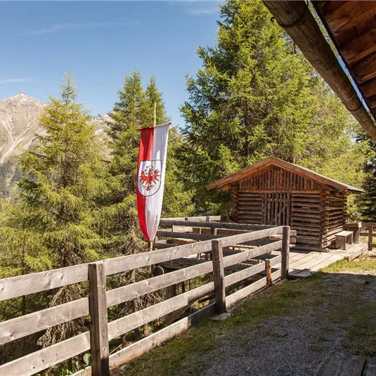 Eine rustikale Berghütte umgeben von hohen Tannen. Im Vordergrund steht eine sichtbare Fahne und eine Holzbank.