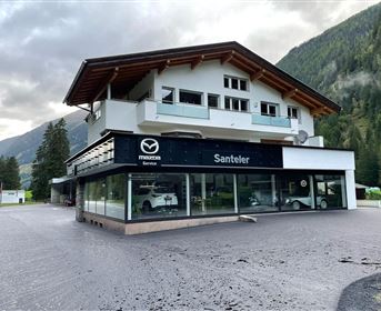 Ein modernes Autohaus mit großen Fensterfronten und dem Logo von Mazda. Im Hintergrund sind Berge und ein bewölkter Himmel sichtbar.