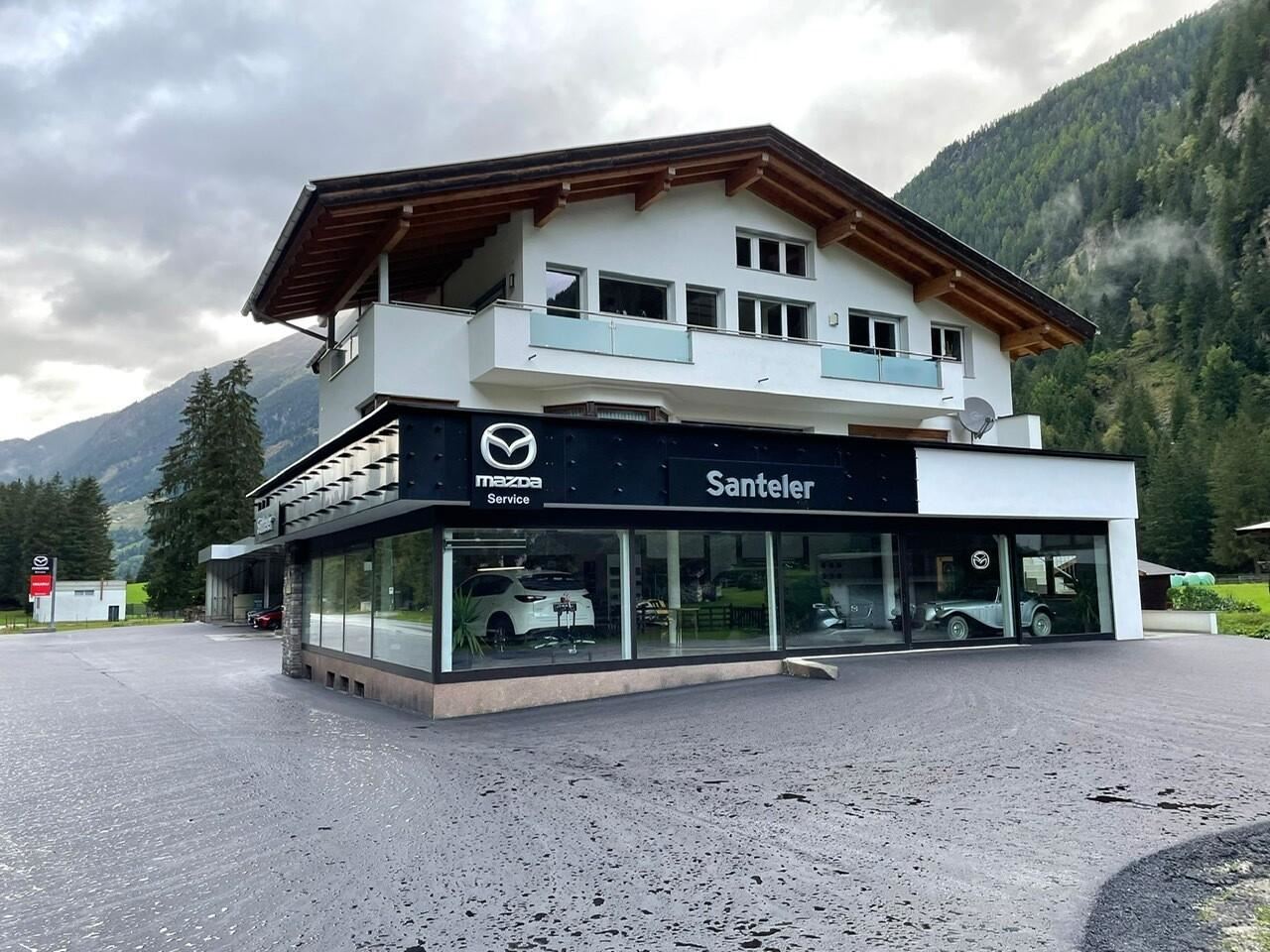 Ein modernes Autohaus mit großen Fensterfronten und dem Logo von Mazda. Im Hintergrund sind Berge und ein bewölkter Himmel sichtbar.