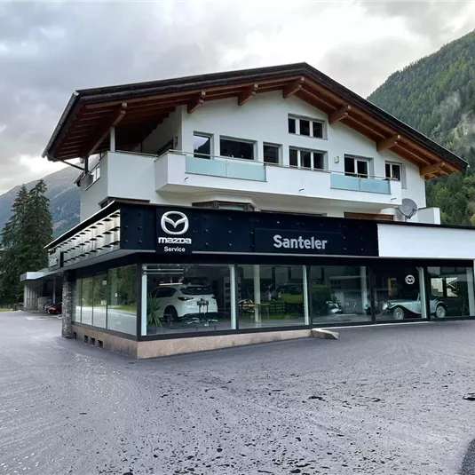 Ein modernes Autohaus mit großen Fensterfronten und dem Logo von Mazda. Im Hintergrund sind Berge und ein bewölkter Himmel sichtbar.
