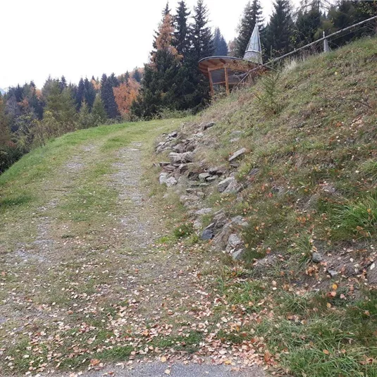 Ein schmaler Weg führt zu einem Hang mit gelegentlichen Steinen. Im Hintergrund sind Bäume und ein Aussichtspunkt zu sehen.