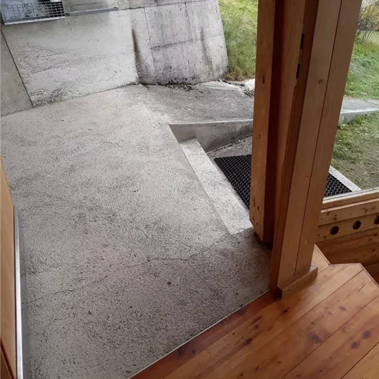 Ein Eingang mit einer Holztreppe und einem Betonboden. Im Hintergrund sind grüne Wiesen und eine Wand sichtbar.