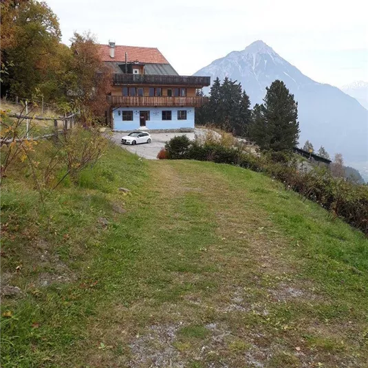Ein ruhiges Haus in den Bergen, umgeben von Bäumen und einer grünen Wiese. Im Hintergrund sind schneebedeckte Berge zu sehen.