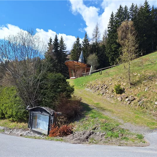 A beautiful, green landscape with a path leading to a small chapel. To the right, there are trees and a clear sky with some clouds.