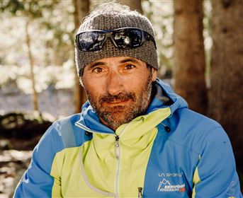 Ein Mann sitzt im Wald mit einer sportlichen Jacke und Sonnenbrille. Der Hintergrund zeigt Bäume und weiches Licht.