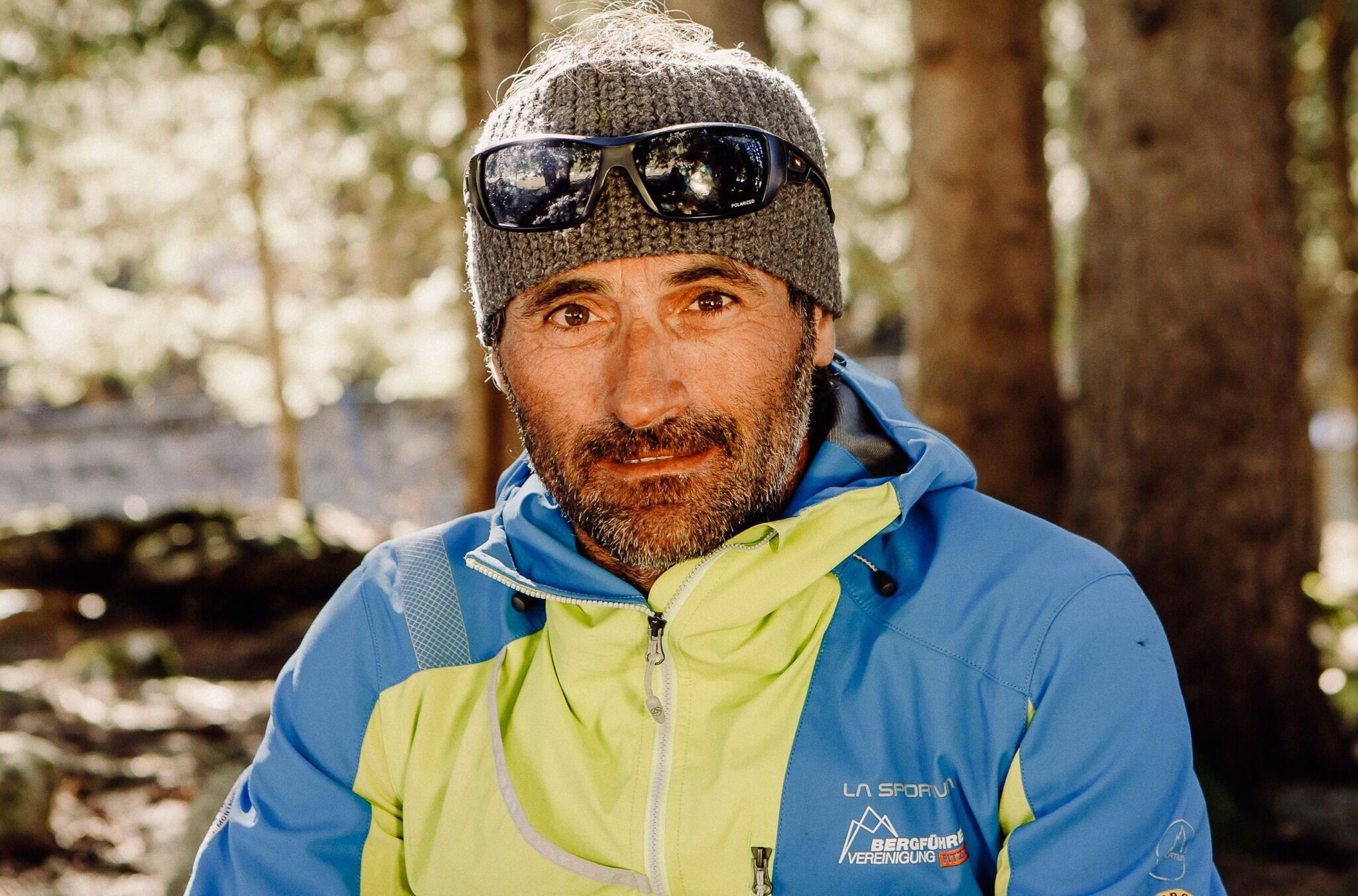 Ein Mann sitzt im Wald mit einer sportlichen Jacke und Sonnenbrille. Der Hintergrund zeigt Bäume und weiches Licht.