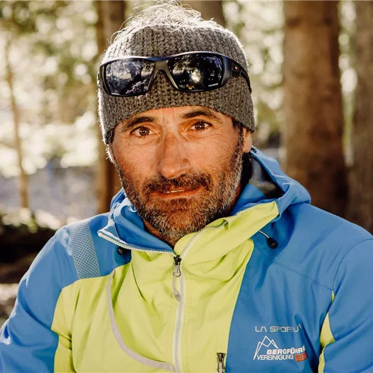 Ein Mann sitzt im Wald mit einer sportlichen Jacke und Sonnenbrille. Der Hintergrund zeigt Bäume und weiches Licht.