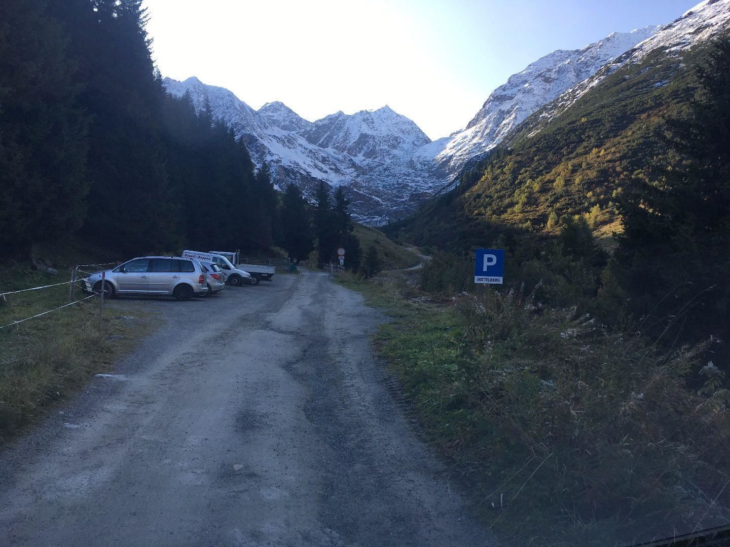 A narrow, unpaved road winds through a mountainous landscape. In the background are snow-capped peaks and a few cars are parked at the roadside.