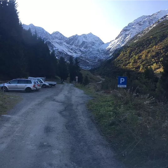 A narrow, unpaved road winds through a mountainous landscape. In the background are snow-capped peaks and a few cars are parked at the roadside.