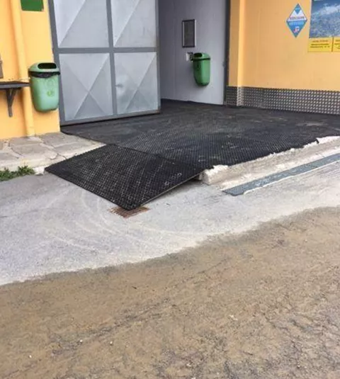 A flat access with a non-slip surface leads to a building. On the left side, there are garbage bins and an entrance with a large door.