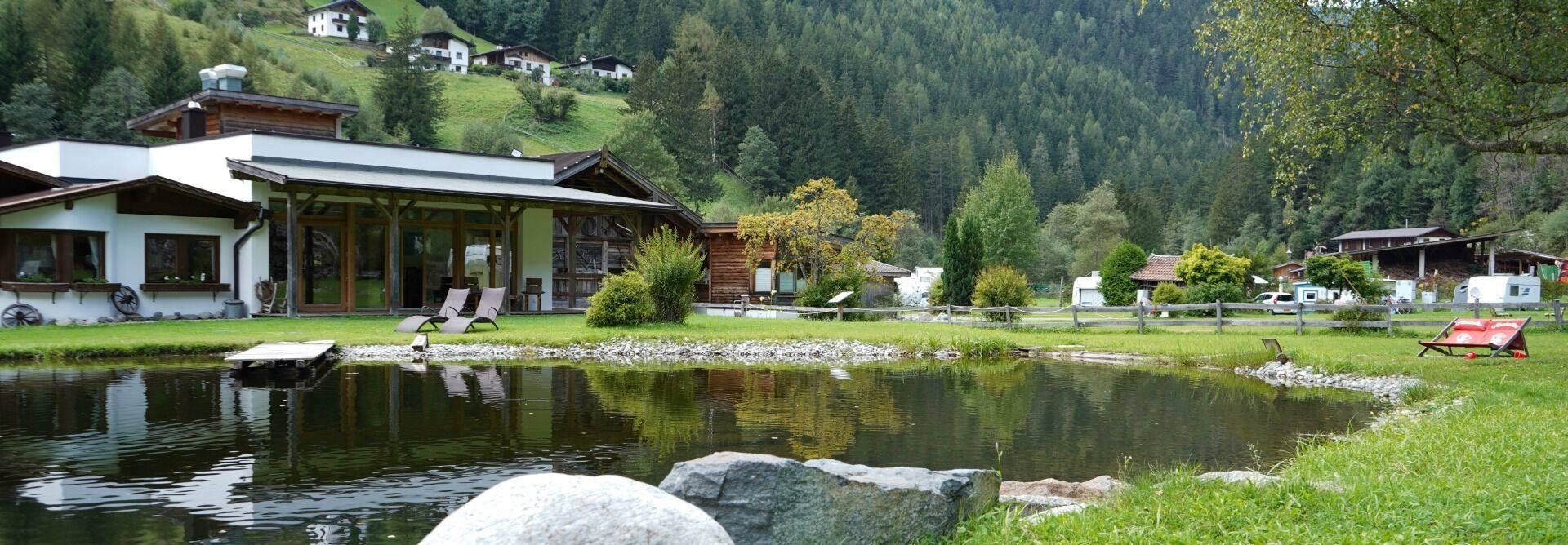 Eine ruhige Landschaft mit einem Teich und einer schönen Aussicht auf die Berge. Im Vordergrund stehen Liegestühle und viel Grünfläche.
