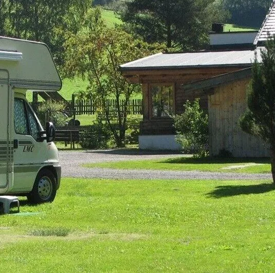 Ein Wohnmobil steht auf einer grünen Wiese vor einem gemütlichen Haus. Im Hintergrund sind Bäume und Sträucher zu sehen.