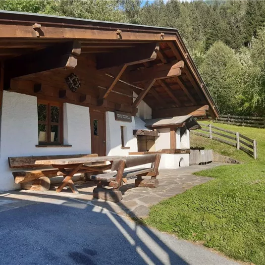Ein traditionelles Holzhaus mit einer Steinbank im Freien. Die Umgebung ist von grünen Wiesen und Bäumen umgeben.