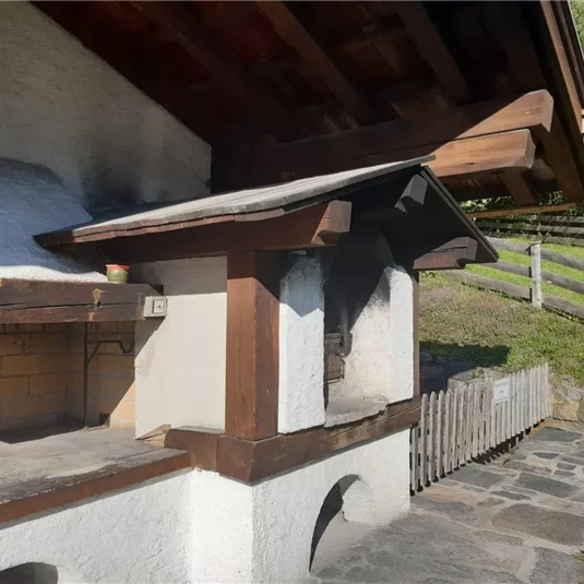 Eine traditionelle Grillstelle aus Holz und Stein in einer ländlichen Umgebung. Im Hintergrund sind Zäune und Bäume sichtbar.