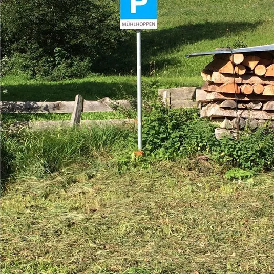 Ein Parkplatzschild mit der Aufschrift "Mühlhoppen" steht neben einem Holzstapel. Im Hintergrund sind grüne Wiesen und Bäume zu sehen.